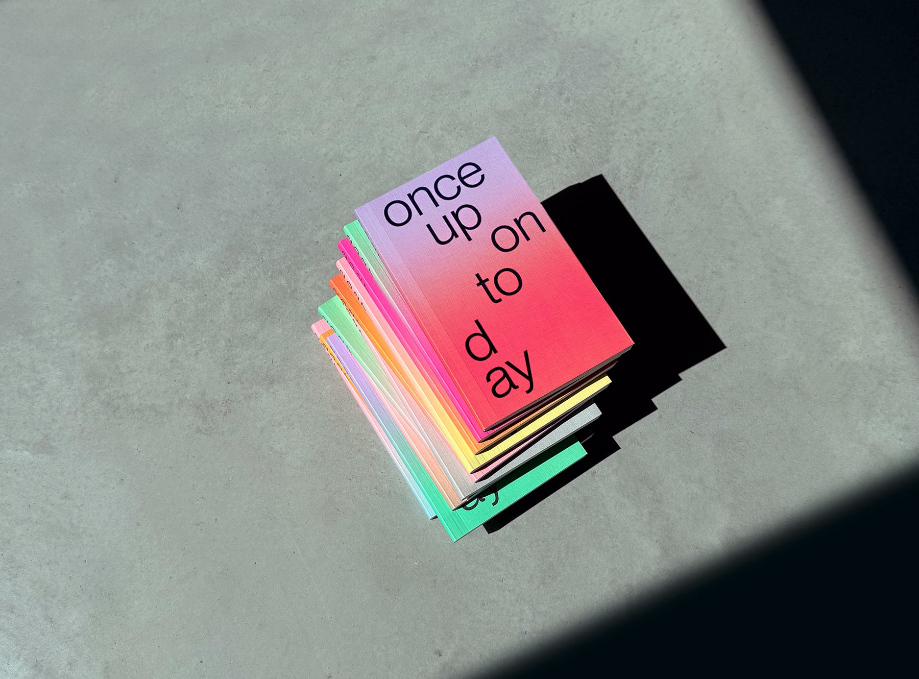 A stacks of edition julie joliat planner 2027 with black text reading 'once upon today' on the top cover, photographed on a concrete surface in dramatic sunlight and shadow.
