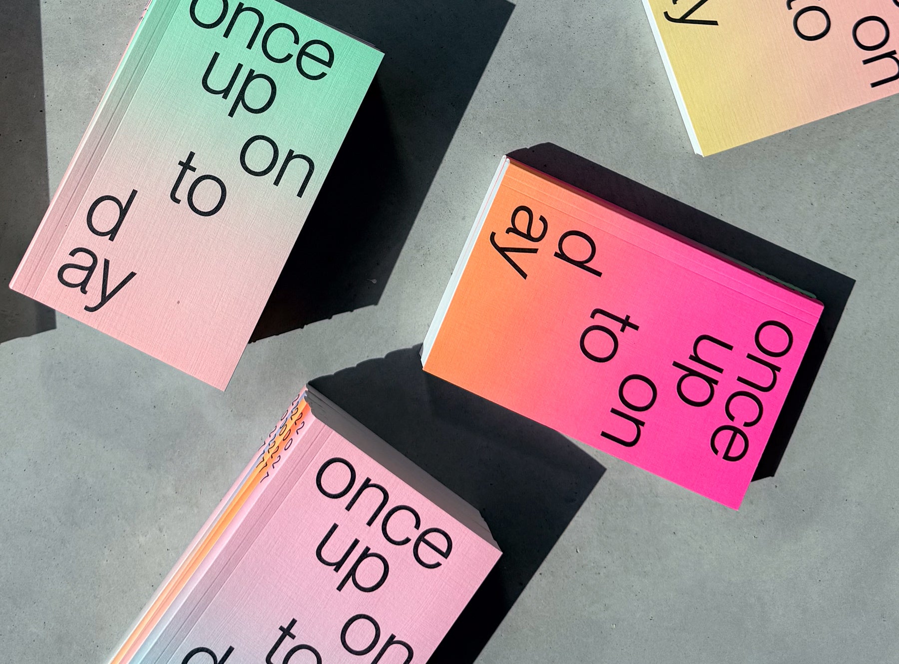 Several stacks of edition julie joliat planner 2027 with black text reading 'once upon today' on the top cover, photographed on a concrete surface in dramatic sunlight and shadow.
