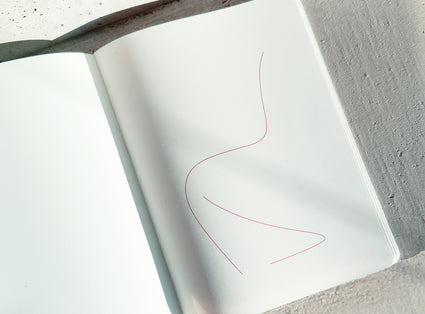 An open white notebook or book lying on a textured gray surface, with a thin pink abstract line drawing on the right page.