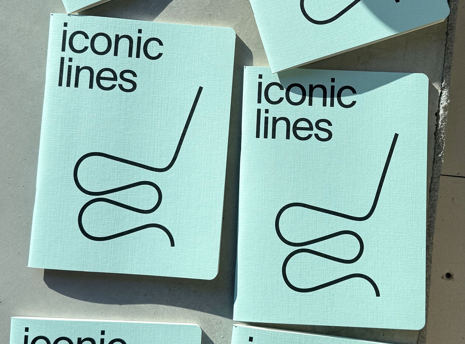 A stack of notebooks with the title 'iconic lines' printed in black sans-serif text and a simple black line drawing on the cover, photographed on a neutral surface.