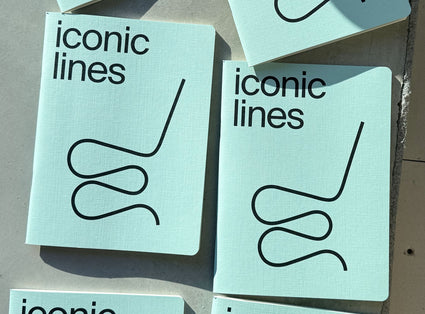 A stack of notebooks with the title 'iconic lines' printed in black sans-serif text and a simple black line drawing on the cover, photographed on a neutral surface.