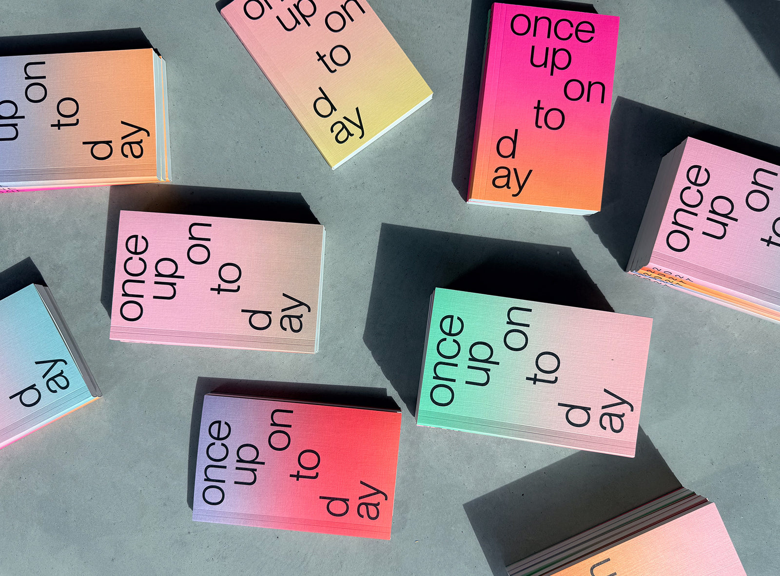 A set of edition julie joliat planner agenda 2027 with a pastel gradient cover and black text reading 'once upon today', arranged on a gray surface with strong sunlight and shadows.