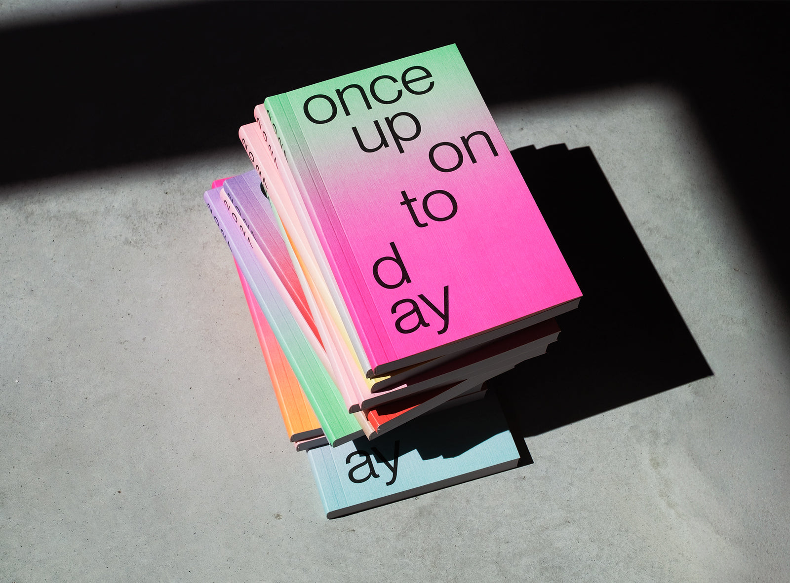 A stack ofedition julie joliat planner agenda 2027 with black text reading 'once upon today' on the top cover, photographed on a concrete surface in dramatic sunlight and shadow.