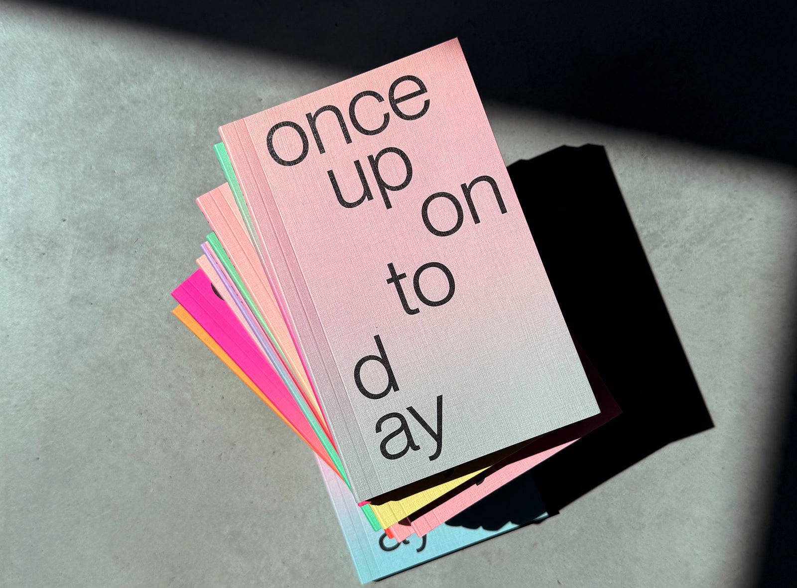 A stack ofedition julie joliat planner agenda 2027 with black text reading 'once upon today' on the top cover, photographed on a concrete surface in dramatic sunlight and shadow.