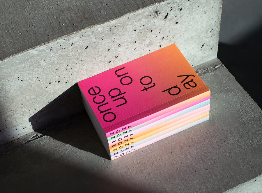 A stack ofedition julie joliat planner agenda 2027 with black text reading 'once upon today' on the top cover, photographed on a concrete stairs in dramatic sunlight and shadow.