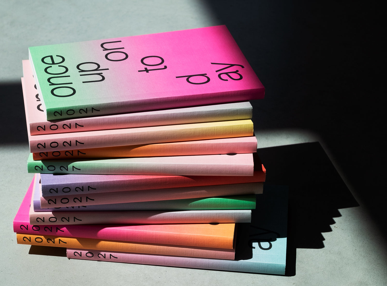 A stack ofedition julie joliat planner agenda 2027 with black text reading 'once upon today' on the top cover, photographed on a concrete surface in dramatic sunlight and shadow.